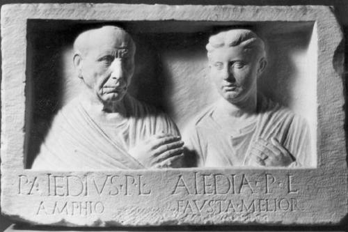 Die Gesichter der Männer – ehemalige Sklaven – sind von Arbeit und Sorge gezeichnet. Das „L“ in der Inschrift steht für „libertus“, Freigelassener. Augusteisches Grabrelief in Berlin, Staatliche Museen.