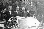 Auf diesen Augenblick hatten die Zuschauer in Dahlem gewartet: John F. Kennedy kommt an – im offenen Wagen mit Berlins Regierendem Bürgermeister, Willi Brandt, und Bundeskanzler Konrad Adenauer