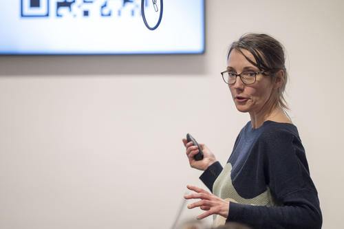Eine Frau mit Brille und dunkelblauem Pullover hält eine Präsentation. Sie spricht und gestikuliert mit einer Hand, während sie in der anderen eine Fernbedienung hält. Im Hintergrund ist ein Bildschirm mit einem QR-Code oder einer Grafik zu sehen.