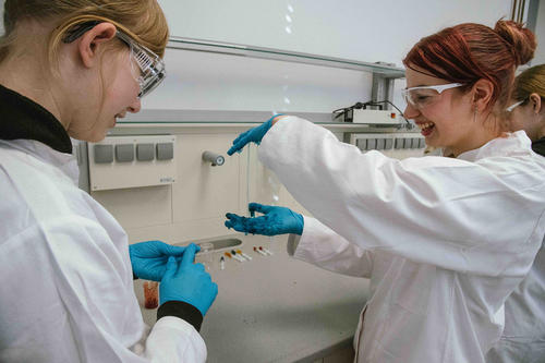 Zwei Mädchen in Laborkitteln und Schutzbrillen experimentieren lachend mit einer schleimartigen Substanz. Beide tragen blaue Schutzhandschuhe, eine hält die Substanz in die Höhe, während die andere interessiert zuschaut.