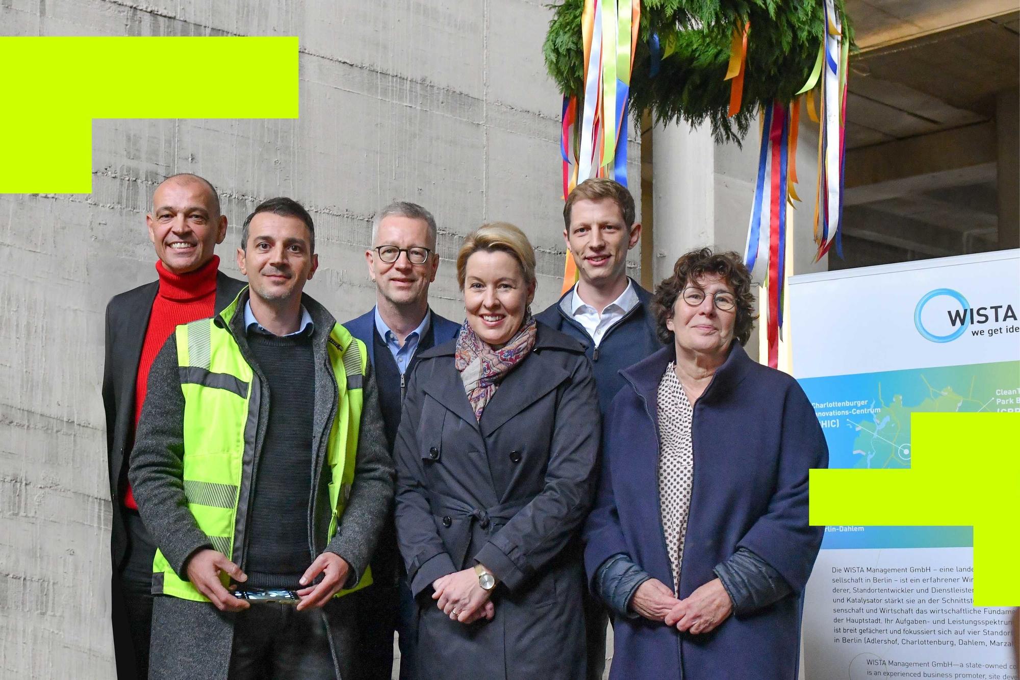 Gruppenfoto von sechs Personen bei einer Baustellenveranstaltung. Im Hintergrund hängen bunte Bänder und ein Richtkranz, daneben ein WISTA-Plakat. Eine Person trägt eine Warnweste, die anderen sind in Businesskleidung.