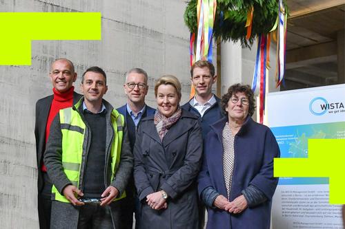 Gruppenfoto von sechs Personen bei einer Baustellenveranstaltung. Im Hintergrund hängen bunte Bänder und ein Richtkranz, daneben ein WISTA-Plakat. Eine Person trägt eine Warnweste, die anderen sind in Businesskleidung.