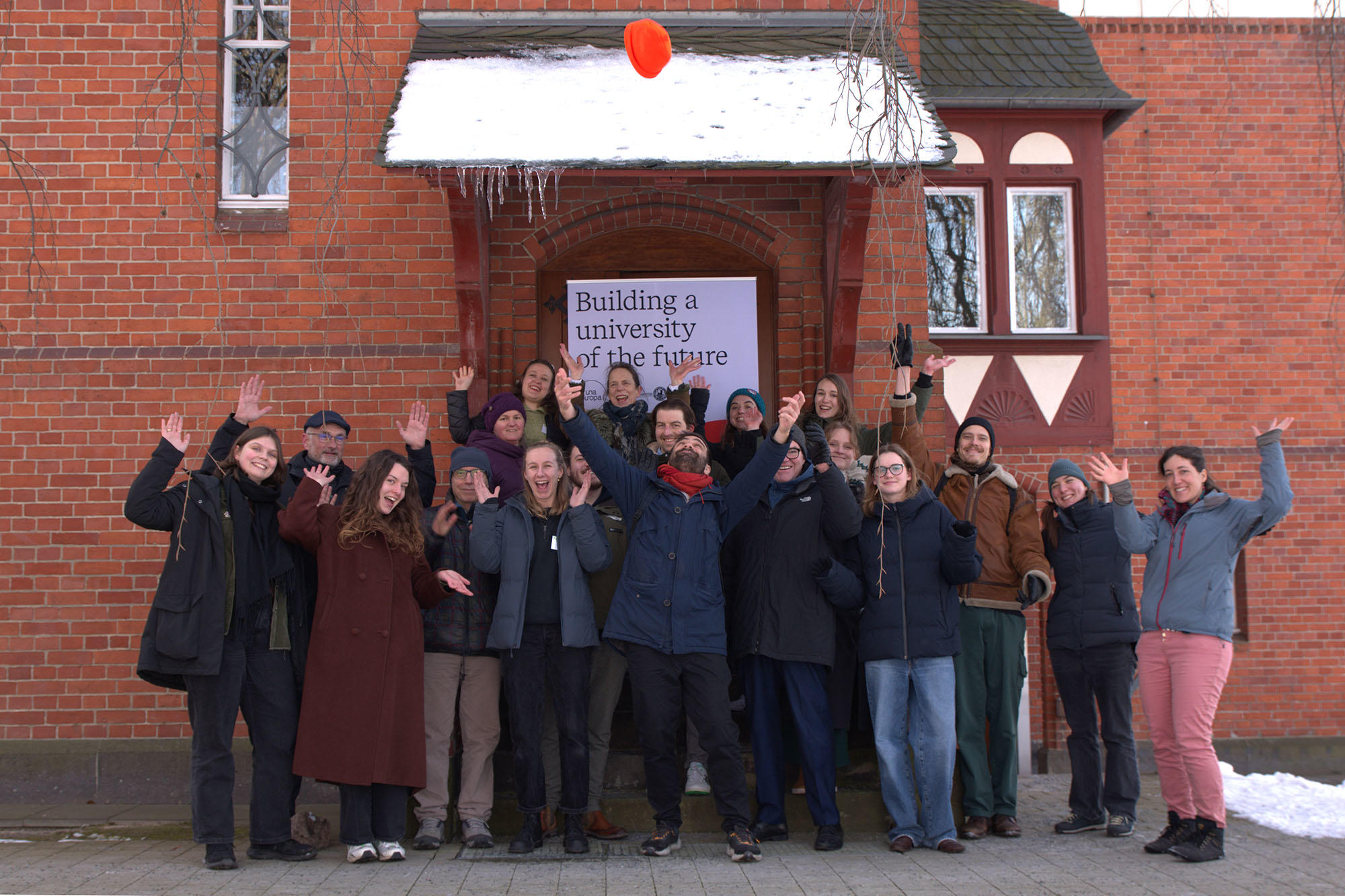 Eine fröhliche Gruppe von Menschen steht vor einem roten Backsteingebäude mit dem Schild „Building a university of the future“. Alle winken oder jubeln, während eine Person in der Mitte eine orangefarbene Mütze in die Luft wirft. Schnee liegt auf dem Dach