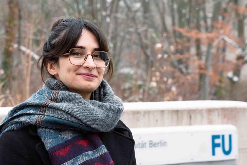 Maryam Keshavarz, dunkle Haaren und Brille, steht im Freien vor einem Schild der Freien Universität Berlin. Sie trägt einen Schal und einen dunklen Mantel. Im Hintergrund sind Bäume mit leichtem Schneefall zu sehen. Sie lächelt und blickt in die Kamera.