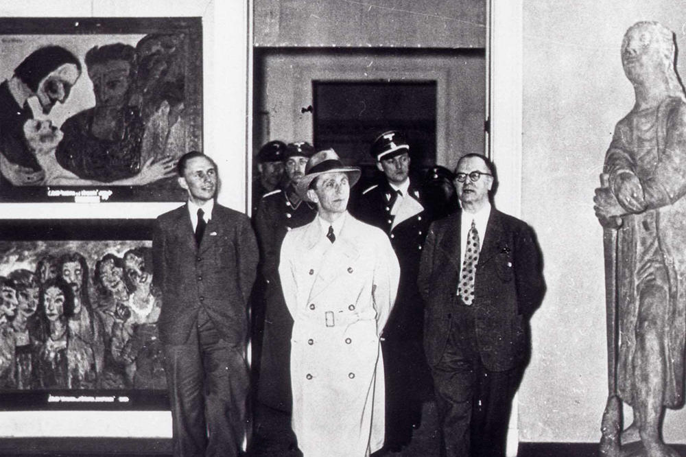 Joseph Goebbels in Mantel und Hut, geht durch eine Tür in den Ausstellungsräumen im Berliner Haus der Kunst, gefolgt von Männern in Uniform. Links von ihm hängen zwei Gemälde, rechts steht eine Skulptur.