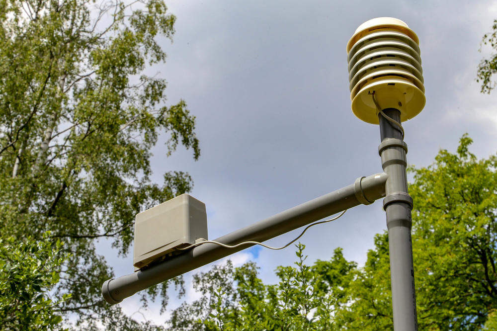 Messgerät aus passiv belüftetem Gerät mit Sensoren für Temperatur, Feuchte, Druck, Strahlung sowie Solarzellen und Akku (re.), Niederschlagsmesser (li.).