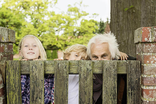 Ein Mädchen, ein Junge und ein älterer Herr blicken hinter einem Gartentor hervor.